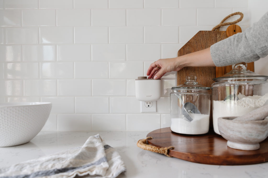 Eine Person wechselt die AERA Mini Düfte im Gerät aus. Das Duftgerät steckt in der Steckdose an einer weißen Wand. Links daneben ist ein Waschbecken. Rechts stehen Mehl, Zucker und ein Mörser auf einem Holzbrett.