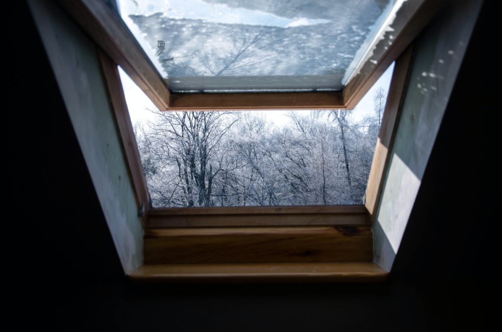 Zu sehen ist en offenes Fenster in einer Dachschrägen. Der Blick geht von drinnen nach draußen auf einen schneebedeckten Wald.